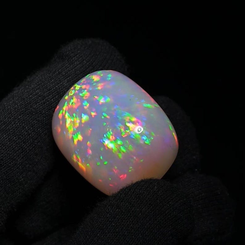 K&ouml;nnte beinhalten: Ein wei&szlig;er Opal-Cabochon mit einem Regenbogen aus irisierenden Farben. Der Opal ist ein nat&uuml;rlicher Edelstein mit einer glatten, polierten Oberfl&auml;che.
