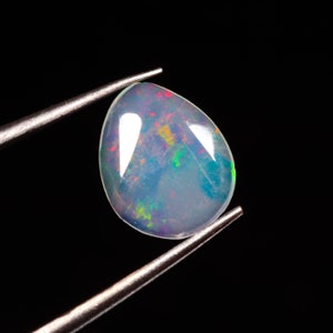 Könnte beinhalten: Ein birnenförmiger Opal-Edelstein, gehalten von einer Pinzette vor schwarzem Hintergrund. Der Opal zeigt ein Farbspiel mit roten, gelben und grünen Blitzen in einer durchscheinenden, milchig-blauen Basis. Der Stein ist poliert und glatt.