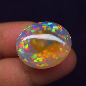 Könnte beinhalten: Ein polierter, ovaler Opal-Edelstein. Der Opal zeigt ein Farbspiel mit grünen, orangefarbenen, blauen und gelben Blitzen. Der Edelstein hat eine glatte, reflektierende Oberfläche und ist vor einem dunklen Hintergrund platziert.