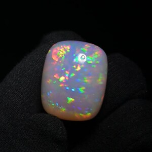 K&ouml;nnte beinhalten: Ein wei&szlig;er Opal-Cabochon mit einem Regenbogen an Farben, darunter Gr&uuml;n, Blau, Rot und Orange. Der Opal ist ein nat&uuml;rlicher Edelstein mit einem einzigartigen und sch&ouml;nen Muster.