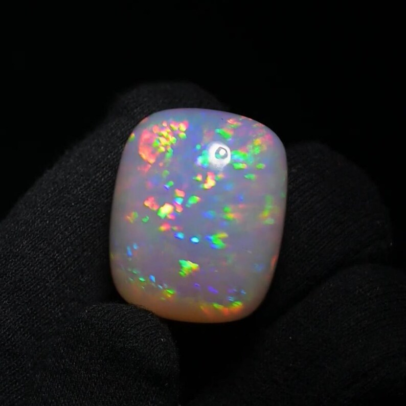 K&ouml;nnte beinhalten: Ein wei&szlig;er Opal-Cabochon mit einem Regenbogen an Farben, darunter Gr&uuml;n, Blau, Rot und Orange. Der Opal ist ein nat&uuml;rlicher Edelstein mit einem einzigartigen und sch&ouml;nen Muster.