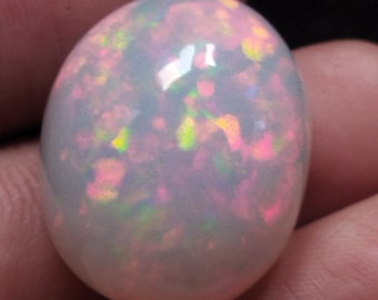May include: A white opal gemstone with iridescent pink, green, and yellow flashes. The opal is oval shaped and has a smooth, polished surface.