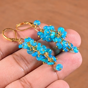 Könnte beinhalten: Ein Paar Ohrhänger mit Clustern aus kleinen, runden, leuchtend blauen Perlen, die an goldfarbenem Metalldraht befestigt sind. Jeder Ohrring hängt an einem goldfarbenen Klappbügel und bildet ein kaskadierendes Design aus vielen Perlen.