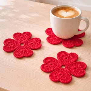 Peut inclure: Une tasse blanche avec un latte art en forme de cœur repose sur un dessous de verre rouge en forme de trèfle crocheté. Deux autres dessous de verre rouges en forme de trèfle sont visibles sur une surface en bois clair. Les dessous de verre sont faits à la main.