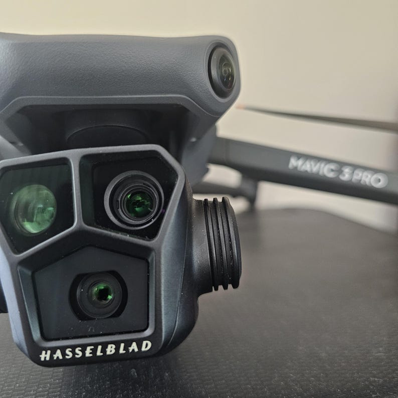 May include: Close-up of a black drone camera with a Hasselblad logo. The camera has three lenses, one large and two smaller. The drone is labeled "MAVIC 3 PRO".