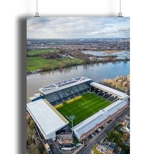 Puede incluir: Vista aérea de un estadio de fútbol con un campo verde y gradas blancas. El estadio está situado en la orilla de un río con un horizonte de ciudad en el fondo.