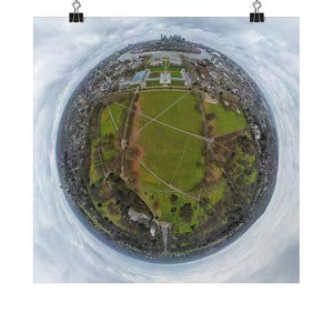 May include: An aerial view of a large green park with a white building in the centre. The park is surrounded by trees and buildings. The sky is cloudy.