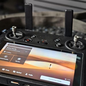 May include: A black drone remote control with a screen displaying a desert landscape and the text "Before You Fly" and "No Fly Spots available nearby". The screen also shows a "Connection Guide" button.