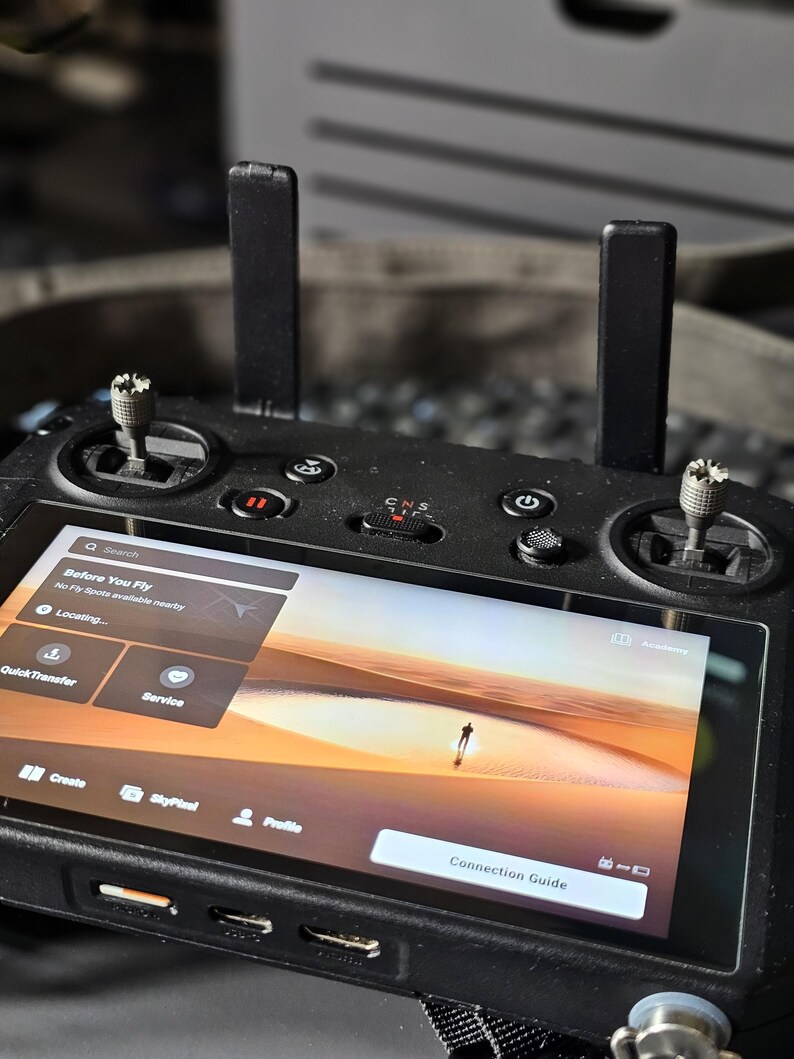 May include: A black drone remote control with a screen displaying a desert landscape and the text "Before You Fly" and "No Fly Spots available nearby". The screen also shows a "Connection Guide" button.