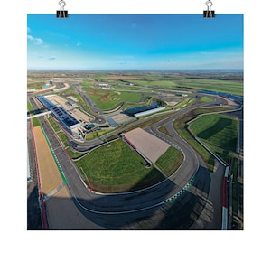 May include: Aerial view of a race track with a long, winding asphalt track surrounded by green grass and a blue sky. The track has several turns and a large, open area in the center.