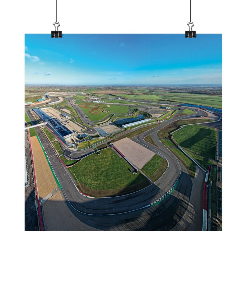 May include: Aerial view of a race track with a long, winding asphalt track surrounded by green grass and a blue sky. The track has several turns and a large, open area in the center.
