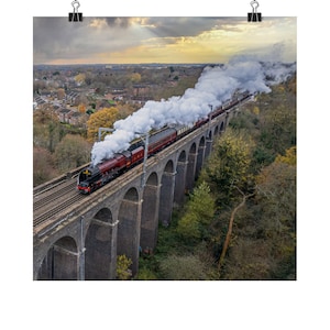 Peut inclure: Un train à vapeur rouge avec des accents noirs traverse un pont de pierre avec une longue traînée de fumée blanche derrière lui. Le train traverse une zone boisée avec une ville au loin.