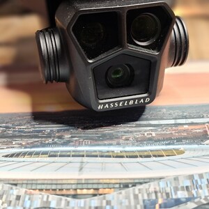 May include: A black camera with three lenses, labeled "HASSELBLAD", is positioned above a printed image of a stadium.