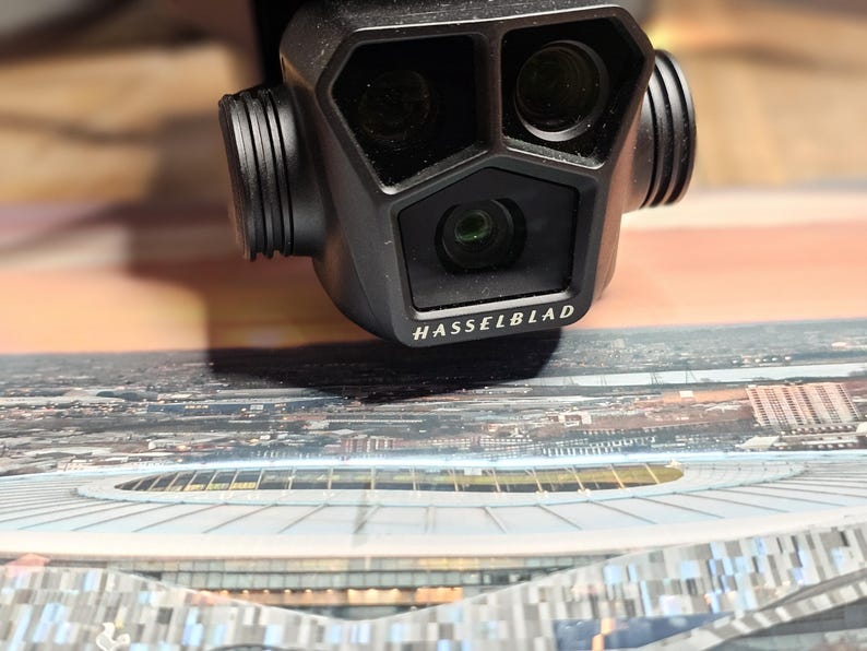 May include: A black camera with three lenses, labeled "HASSELBLAD", is positioned above a printed image of a stadium.