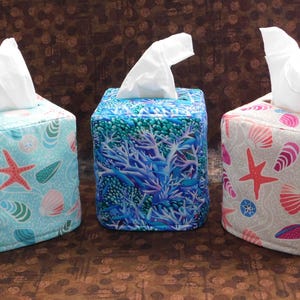 May include: Three decorative tissue box covers with coastal themes. One is light blue with starfish and seashells, another is blue with coral, and the third is off-white with pink and red seashells and starfish. Each box has white tissues emerging from the top.