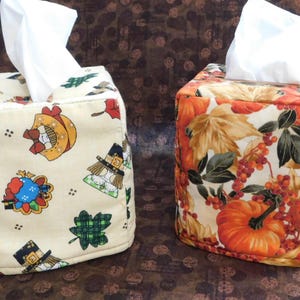 May include: Two fabric tissue box covers. One is beige with fall-themed patterns, including pumpkins and pilgrims. The other is orange with fall leaves and pumpkins. Both have white tissues emerging from the top.