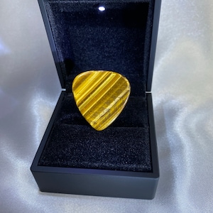 Puede incluir: Una púa de guitarra de ojo de tigre dorada y marrón en una caja negra forrada de terciopelo con una luz en el interior.