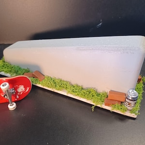 Peut inclure: Un obstacle miniature de type rebord en béton pour fingerboard, comprenant un long bloc gris sur une base décorée de mousse verte, de petites briques rectangulaires brunes et une minuscule canette, avec un finger skateboard rouge.