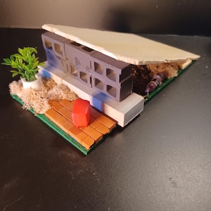 Puede incluir: Una maqueta arquitectónica en miniatura con una estructura de bloques de hormigón gris y un tejado de color claro. El modelo incluye una pequeña planta verde en una maceta blanca, un cubo rojo y una cubierta de madera. La base es verde con musgo beige.