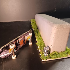 Puede incluir: Un fingerboard con tabla gráfica y ejes plateados se sitúa junto a un obstáculo de tipo repisa de hormigón. La repisa está sobre una base rectangular cubierta de musgo verde, con una lata de refresco en miniatura con texto coreano.