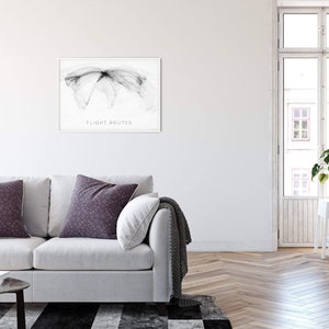 May include: A white sofa with two throw pillows and a gray throw blanket in a living room with a framed black and white print of flight routes on the wall. The room has a white painted wall, a wood floor, and a window with a view of a building.