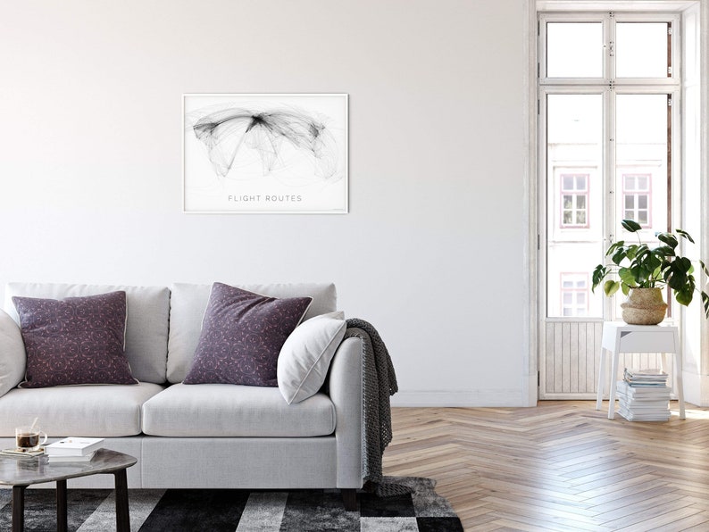 May include: A white sofa with two throw pillows and a gray throw blanket in a living room with a framed black and white print of flight routes on the wall. The room has a white painted wall, a wood floor, and a window with a view of a building.