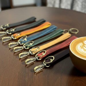 May include: A collection of seven leather wristlet keychains in various colors including black, orange, blue, green, yellow, red, and dark brown. Each keychain features a flat leather strap, a brass-toned metal lobster clasp, and a split ring.