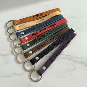 May include: A collection of leather keychains in various colors, including tan, blue, red, and purple. Each keychain has a metal ring and is embossed with phrases like "I Love You Mom" and "Drive Safe."
