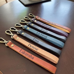 May include: Six leather keychains in various colors including tan, light brown, blue, dark blue, purple, and orange. Each strap features a metal lobster clasp, a gold-tone rivet, and a name engraved in black: Lakeesha, Denise, Tina, La Nesa, Paula, Yolanda, and Patricia.