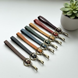 May include: A collection of eight leather wristlet keychains arranged in a diagonal row, featuring various colors including brown, black, tan, blue, green, yellow, and red. Each strap includes a brass-toned metal split ring and a lobster clasp.