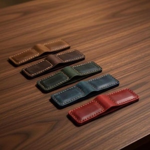 May include: Five leather money clips in various colors: brown, dark brown, green, blue, and red. Each clip is rectangular with a curved center section and white stitching around the edges. The clips are arranged diagonally on a wooden surface.