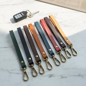 May include: A collection of leather wrist straps in various colors, including green, black, brown, orange, blue, red, and tan. Each strap is attached to a gold-colored clasp. A car key and key fob are in the background.