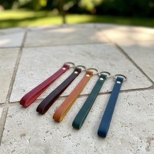 May include: Five leather wristlet keychains arranged in a row, featuring colors of red, dark brown, tan, green, and blue. Each strap is secured with a silver-toned metal rivet and includes a circular metal split ring for attaching keys.
