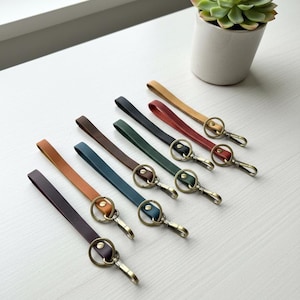 May include: A collection of leather keychains in various colors, including brown, tan, green, and burgundy. Each keychain features a metal ring and clasp for attaching keys. The keychains are arranged on a white surface.
