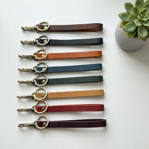 May include: A collection of nine leather wristlet keychains in various colors including dark brown, black, tan, navy blue, forest green, light brown, red, and deep purple. Each keychain features a brass-toned metal lobster clasp and a rivet.