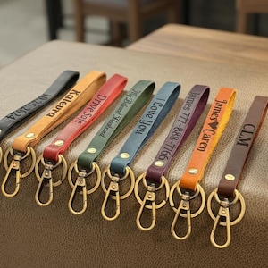 May include: A collection of personalized leather keychains in various colors, including black, orange, red, and green. Each keychain features a gold-colored clasp and is engraved with text such as names, dates, and phrases.