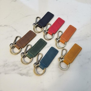 May include: A collection of leather keychains in various colors, including brown, black, red, orange, green, and blue. Each keychain features a rectangular leather tag with a metal clasp and key ring.