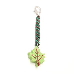 May include: A keychain featuring a light green maple leaf charm with a dark brown vein pattern, attached to a braided cord in alternating dark brown and forest green colors, finished with a silver-toned metal lobster claw clasp at the top.