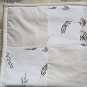 May include: A white quilted baby play mat with a square design. Each square features a different pattern, including a goose with a hat, leaves, and a simple white background.