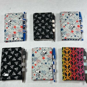 May include: Six different fabric-covered notebooks with pens attached. The notebooks feature various patterns, including a black and white pattern with triangles, a gray and white pattern with stars and fireworks, a gray and white pattern with Snoopy, a gray and white pattern with peace signs, and a black and white pattern with Harry Potter symbols.