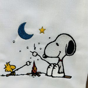 以下が含まれることがあります： 白い布製のタオルに、黒い耳の白い犬と黄色い鳥が夜の小さな焚き火のそばに座っている刺繍が施されています。青い三日月、黄色い星、そしてマシュマロを焼いている両キャラクターが描かれたデザインです。
