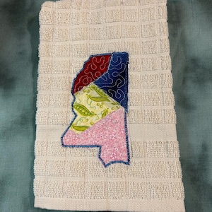 May include: A cream-colored waffle-weave kitchen towel featuring a fabric applique in the shape of the state of Mississippi. The applique is divided into four sections with red, dark blue, green pea-patterned, and pink floral fabric pieces.