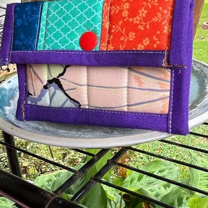 May include: A rectangular quilted fabric clutch featuring a patchwork flap with blue, teal, and orange floral and geometric patterns. The main body has a light-colored panel with a dark abstract design, all bordered by purple fabric and a red button.