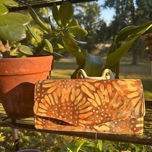 May include: A rectangular wallet featuring a batik-style pattern in shades of brown, orange, and yellow. The exterior displays a repeating floral or leaf-like motif with dotted accents and a flap closure that spans the width of the item.