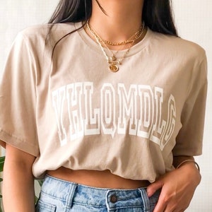 May include: A beige t-shirt with a white graphic that says "YHLQMDLG" in a bold, blocky font.