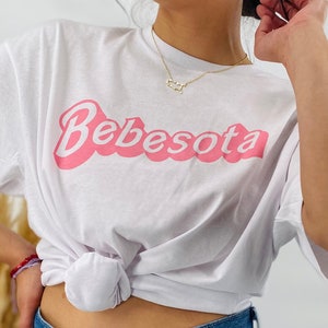 May include: A white t-shirt with a pink graphic that says "Bebesota" in a retro font. The shirt is tied at the waist and the person is wearing blue jeans.