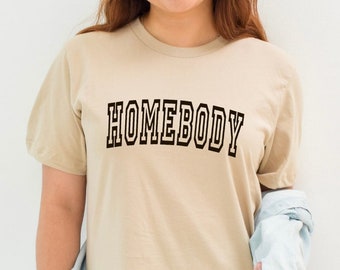 May include: A beige t-shirt with the word "HOMEBODY" printed in black outlined letters.