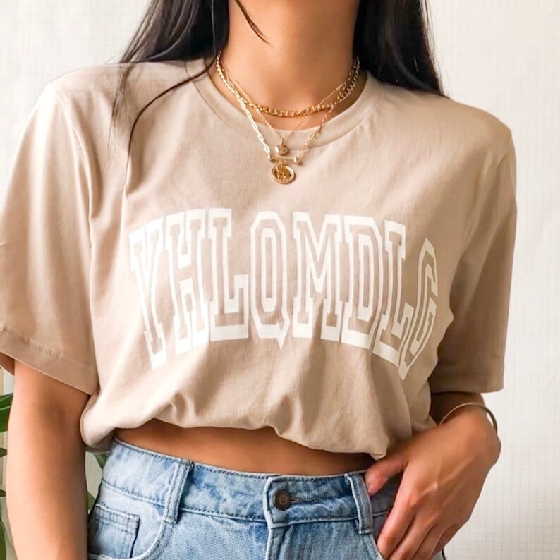 May include: A beige t-shirt with a white graphic that says "YHLQMDLG" in a bold, blocky font.
