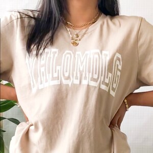 May include: A beige t-shirt with a white graphic that says "YHLQMDLG" in a bold, blocky font.