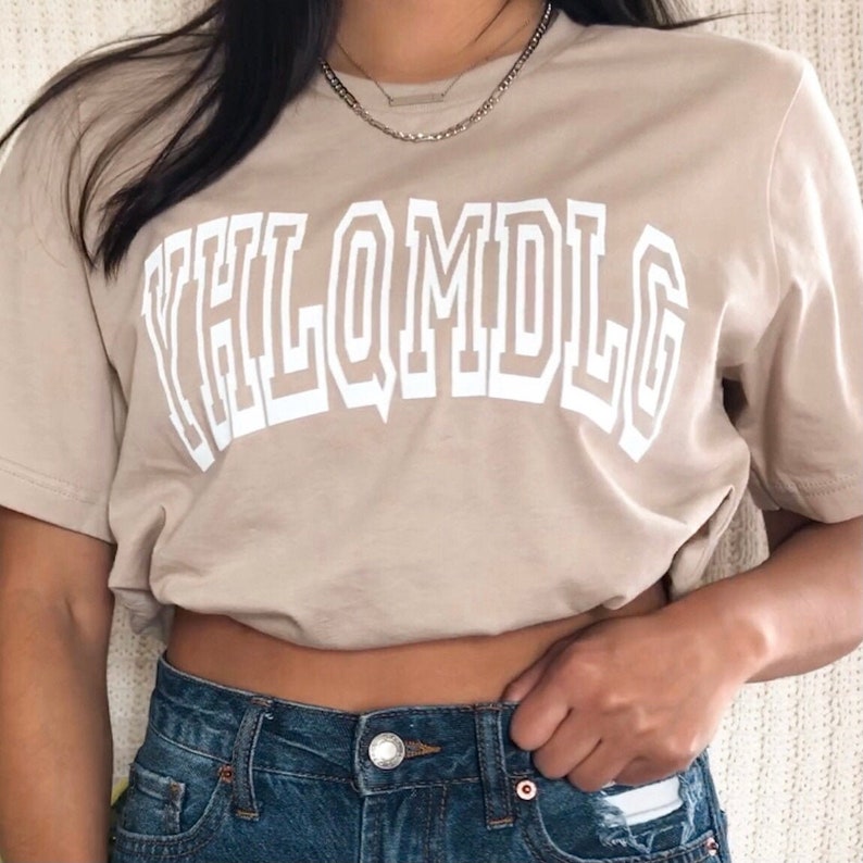 May include: A beige t-shirt with a white graphic that says "YHLQMDLG" in a bold, blocky font.